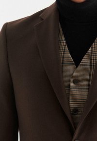 Giacca marrone su misura con revers a punta, indossata sopra un dolcevita nero e un gilet a quadri con motivi neri, marroni e crema.