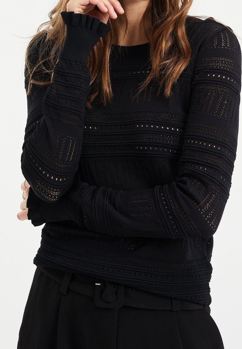 Femme portant un haut noir en maille texturée à manches longues avec des motifs transparents, un pantalon noir et sa main reposant près de son visage. Vue du haut du corps.