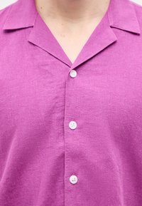 Κοντινό πλάνο ενός ατόμου που φοράει μοβ πουκάμισο με γιακά με εγκοπή και τρία ορατά λευκά κουμπιά.