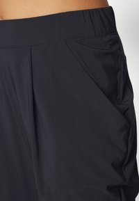 Close-up van een zwarte broek met elastische tailleband en zijzak, gedragen ter hoogte van de taille.