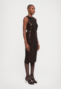 Mujer de pie con un vestido negro de lentejuelas sin mangas y de longitud a la rodilla, medias negras y sandalias de tacón alto y tiras negras, sobre un fondo blanco liso.