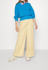 Femme portant un pull bleu vif, un pantalon beige à jambes larges, des chaussures blanches sans lacets et une écharpe colorée autour du cou, debout devant un fond blanc uni.