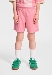 Rosa träningsshorts med en slät textur, elastisk midja och vitt logotyp. Tillsammans med gröna sneakers och vita strumpor.