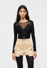 Νεαρή γυναίκα με μακριά σκούρα μαλλιά που φοράει μαύρο διάφανο μακρυμάνικο τοπ και μπεζ σορτς ψηλόμεσο με μεγάλες τσέπες και μαύρο καλσόν.