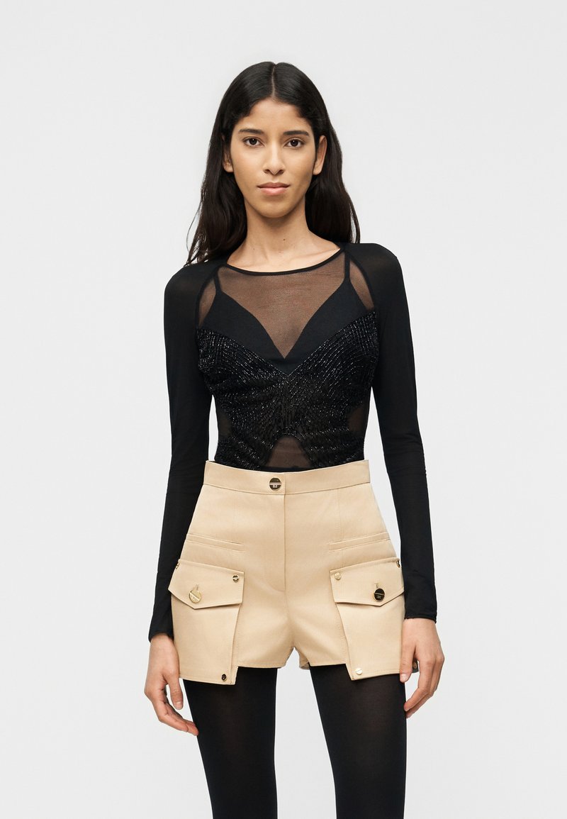 Νεαρή γυναίκα με μακριά σκούρα μαλλιά που φοράει μαύρο διάφανο μακρυμάνικο τοπ και μπεζ σορτς ψηλόμεσο με μεγάλες τσέπες και μαύρο καλσόν.