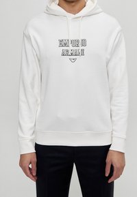 Felpa bianca con il logo "Emporio Armani" in nero, cappuccio con cordino, tessuto morbido e polsini e orlo a costine, indossata con pantaloni scuri.