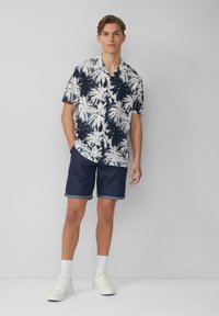 Korte mouwen overhemd met een marineblauw en witte palmboomprint, gecombineerd met donkere spijkerbroekshorts en witte sneakers.