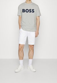Camiseta de algodón gris con gran logotipo negro de "BOSS", combinada con pantalones cortos de algodón blancos. El modelo lleva zapatillas blancas y calcetines hasta el tobillo.