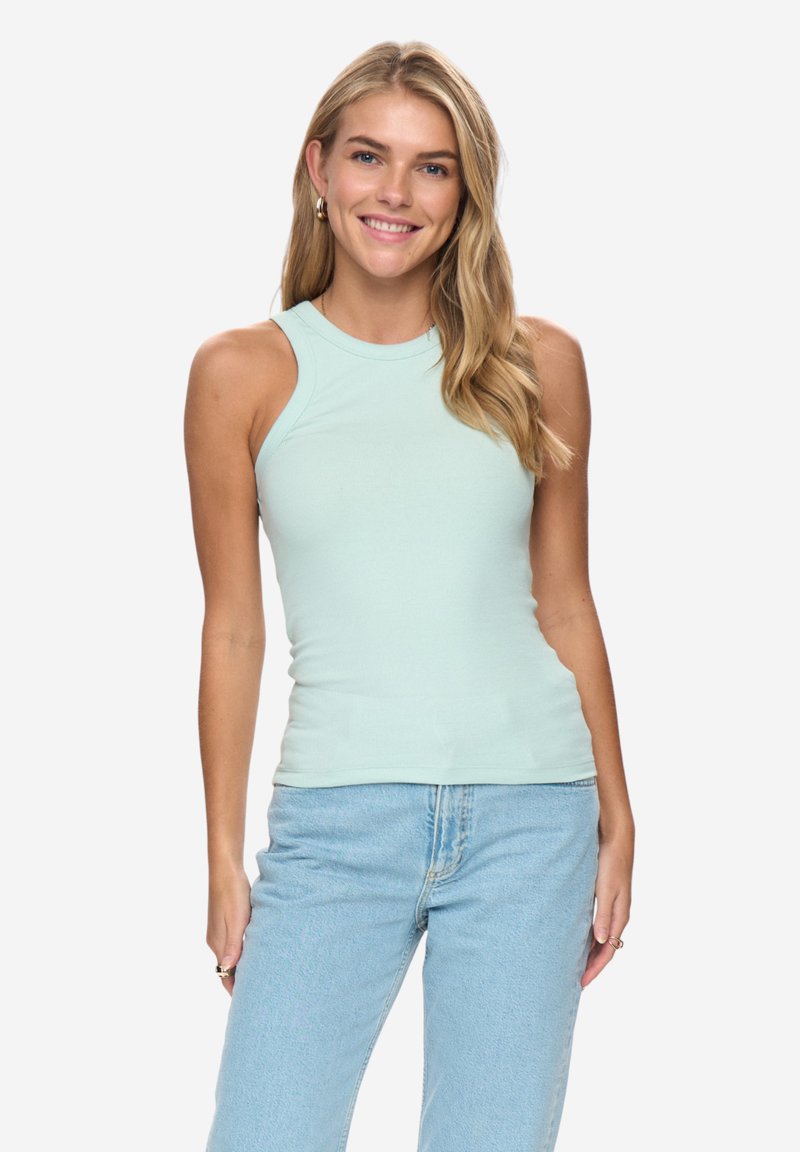 Jeune femme souriante, portant un top sans manches bleu clair et un jean bleu clair, debout devant un fond blanc uni.