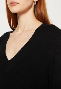 Schwarzer Strickpullover mit V-Ausschnitt, der über eine gerippte Textur verfügt. Nahaufnahme zeigt die Details des Ausschnitts und des Materials.