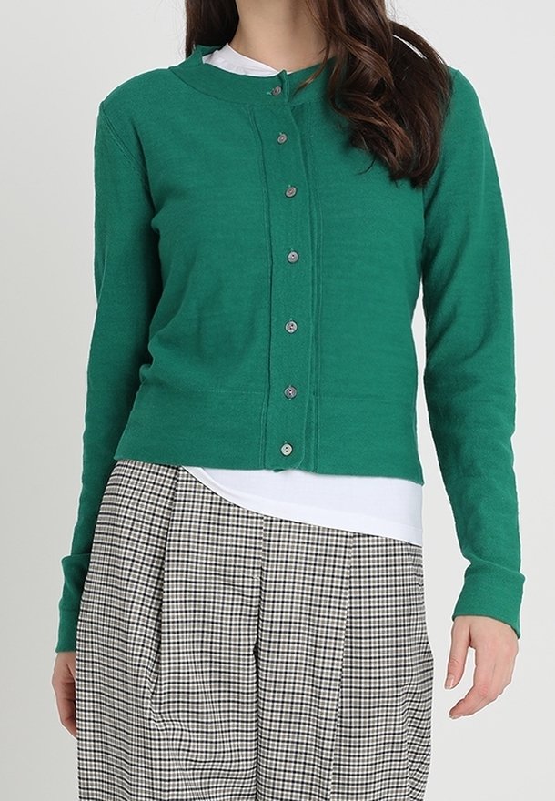Cardigan boutonné vert avec un col arrondi, confectionné dans un tissu doux, avec des manches longues et une texture lisse. Porté par-dessus un haut blanc.
