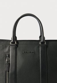 Černá kabelka z texturované kůže s dvěma horními uchy, zipovým uzavřením a embosovaným logem. Má strukturovaný design a hladké akcenty.