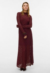 Viininpunainen pitsimallinen maksimekko, jossa on pitkät hihat ja korkea kaulus. Mekossa on läpikuultava kangas ja hienovarainen kuviointi. Yhdistetty teräväkärkisiin korkokenkiin.