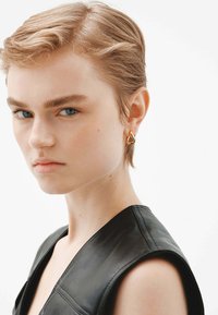 Pendiente de aro de oro con un diseño liso y con forma. El modelo lleva una parte de arriba de cuero negro, con una silueta minimalista y estructurada.