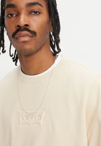 Beige hettegenser med en teksturert "Levi's"-logo. Har en rund hals, en subtil glans, og er lagt over en hvit T-skjorte. Sølv kjede.