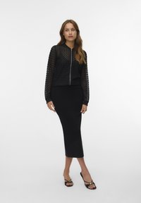Black mesh zip-up bomber jacket with long sleeves over a fitted black midi skirt. Model wearing black strappy sandals. Simple, sleek design.
