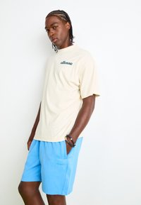 Tricou din bumbac bej cu logo-ul verde "ellesse", asortat cu pantaloni scurți albaștri. Ținuta are o croială lejeră și mâneci scurte.