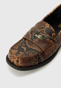 Mocassim em estampado de cobra castanha com sola preta, apresentando uma tira com logotipo na parte superior. Textura suave com um brilho subtil.