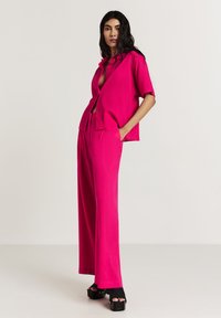 Donna in piedi con le mani nelle tasche, che indossa pantaloni larghi di un brillante colore rosa e una camicia a maniche corte con bottoni, abbinata a sandali con platform neri.