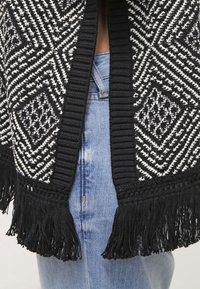 Cardigan en tricot à motif noir et blanc avec des détails en frange à l'ourlet et une bordure côtelée le long de l'ouverture avant.