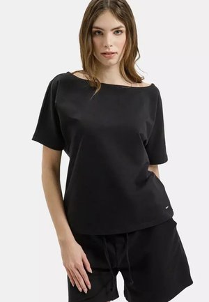 Młoda kobieta o jasnobrązowych włosach, ubrana w luźną czarną bluzkę z krótkim rękawem i dopasowane czarne szorty, stojąca na jednolitym białym tle.