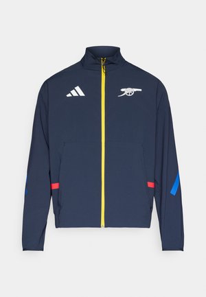 Veste zippée marine en matière légère ; comporte un logo blanc et des accents rouges et bleus, avec une fermeture éclair jaune et un col montant.