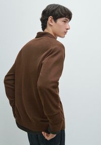 Brauner Strickpullover mit geripptem Kragen und Bündchen, der eine lockere Passform und eine strukturierte Stoffstruktur aufweist. Getragen über einem dunklen Hemd und Jeans.