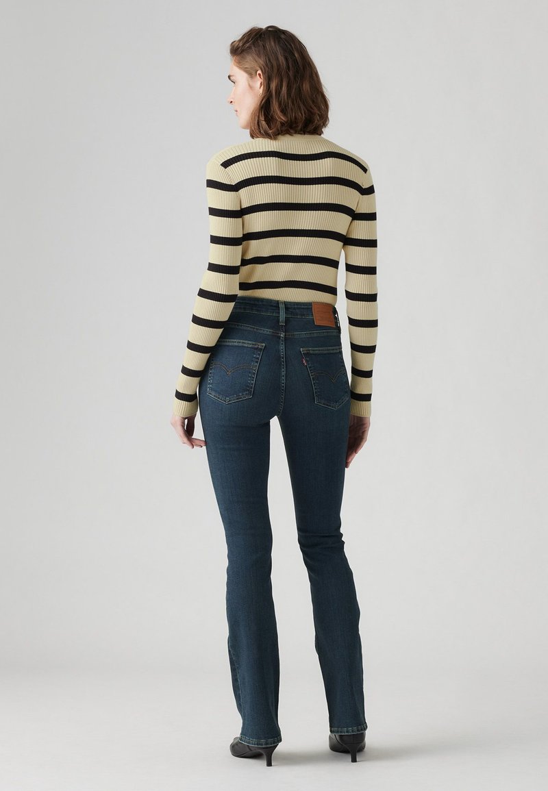 Suéter de rayas acanaladas en amarillo y negro, combinado con vaqueros de campana en azul oscuro que tienen bolsillos traseros y una etiqueta de cuero marrón.