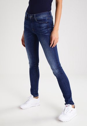 Vrouw draagt donkerblauwe strakke spijkerbroek en witte sneakers, staand met licht gespreide benen tegen een effen witte achtergrond.