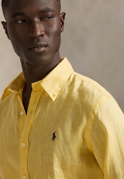 Κίτρινο λινό πουκάμισο με κλασικό γιακά, εμπρόσθιο κούμπωμα με κουμπιά και μικρό μαύρο έμβλημα λογότυπου στο αριστερό στήθος.