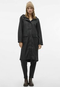 Juodas lietpaltis su gaubtu, spaustomis sagtimis, dviem priekinėmis kišenėmis ir lygia tekstūra. Derinamas su juodomis kelnėmis ir rudos spalvos batais.