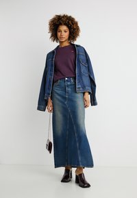 Γυναίκα με σγουρά μαλλιά που φοράει μωβ μπλούζα, φαρδιά τζιν μπουφάν, μακριά τζιν φούστα, μαύρα παπούτσια με τακούνι και κρατάει μια μικρή τσάντα.