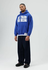 Blauwe hoodie met capuchon en witte letters "HE TRAINS FOR BOXING," voorzak, gecombineerd met donkere, wijde jeans en zwarte schoenen.