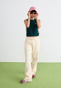 Zöld kötött ujjatlan cropped top, krém színű bársonyos melegítőnadrág, rózsaszín sapka és napszemüveg, sötét vörös és fehér sportcipő. Egyszerű háttér.