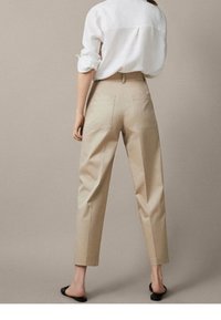 Pantalons taillés beiges à taille haute, dotés de poches latérales et d'une finition lisse. Portés avec une chemise blanche ample et des mules noires.