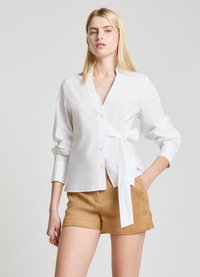 White, button-up shirt with a wrap tie at the waist, long sleeves with cuffs. Paired with tan shorts. Smooth fabric texture.