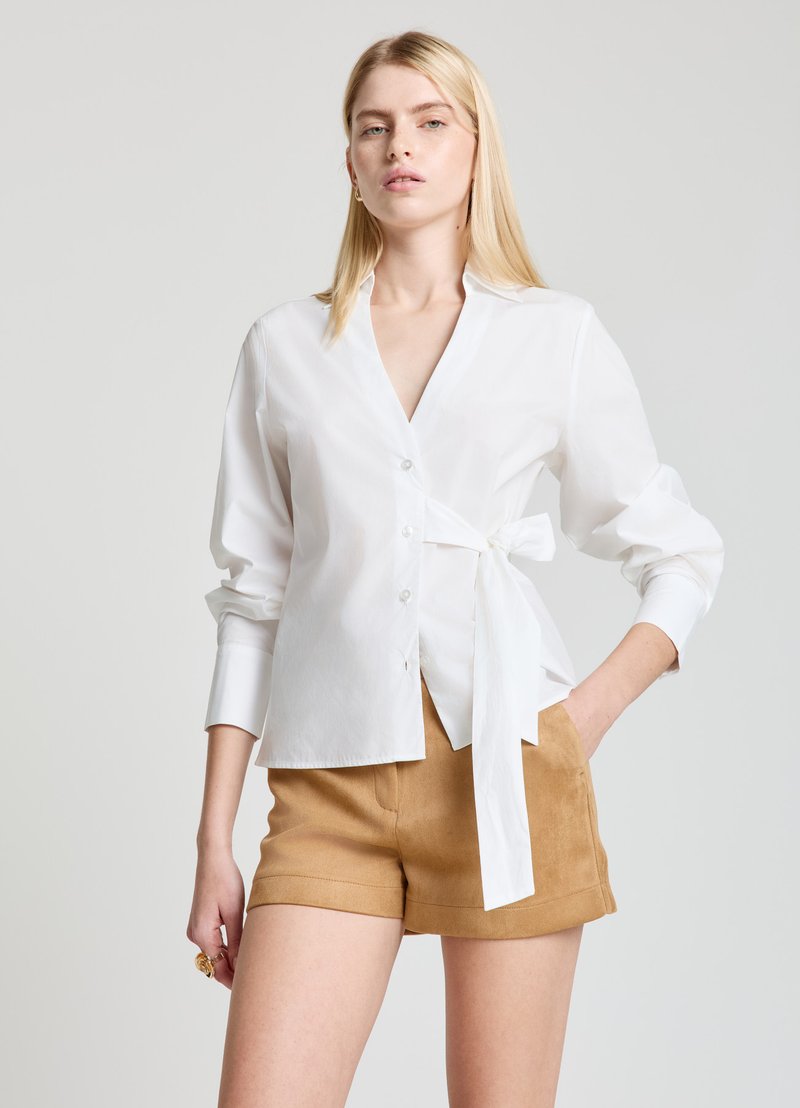 White, button-up shirt with a wrap tie at the waist, long sleeves with cuffs. Paired with tan shorts. Smooth fabric texture.