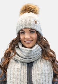 Bonnet tricoté gris avec un pompon en fourrure et une étiquette en cuir beige, assorti à une écharpe à côtes grise. Les deux sont texturés et confortables.