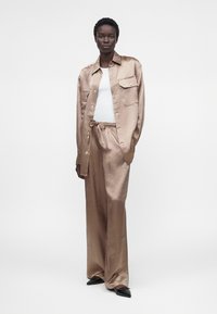 Γυναίκα φοράει μια λαμπερή μπεζ oversized πουκάμισο και αντίστοιχες φαρδιές παντελόνες πάνω από λευκό τοπ, στέκεται μπροστά σε απλό ανοιχτό φόντο.