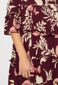 Burgundy blommig klänning med krämfärgade botaniska mönster, texturerad tyg, volangärmar och utställda lager. Hand som håller tyget vid midjan.