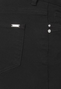 Jean en denim noir avec une étiquette logo argentée sur la poche arrière, des rivets et une texture lisse. Coupe ajustée avec des surpiqûres classiques.