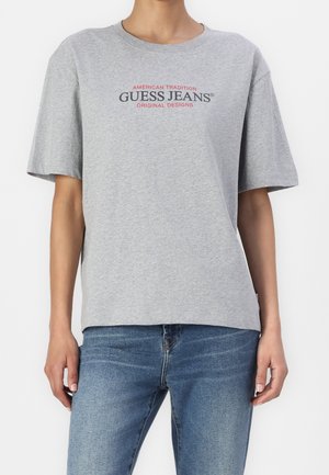 Osoba ubrana w szarą koszulkę z krótkim rękawem z napisem "GUESS JEANS American Tradition Original Designs", w połączeniu z niebieskimi dżinsami.