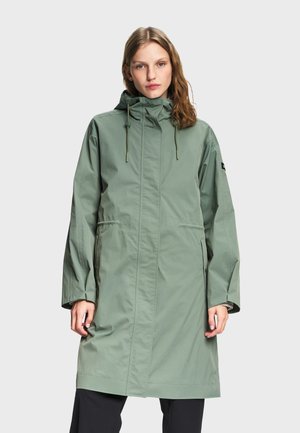 Femme aux cheveux ondulés portant un long imperméable à capuche vert clair sur un pantalon noir, debout devant un fond gris clair uni.