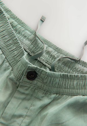 Shorts din bumbac verde deschis, cu un brâu elastic, închidere cu buton negru și șnur. Țesătura are o textură moale și netedă.