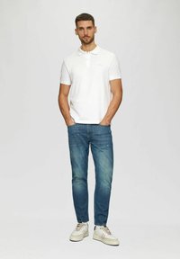 Weißes Polohemd, blaue Jeans und weiße Turnschuhe. Das Polohemd hat einen Kragen und kurze Ärmel; die Jeans haben eine klassische Passform mit leichter Verfärbung.