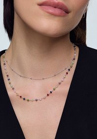Warstwowe naszyjniki z srebrnych łańcuszków ozdobionych wielokolorowymi koralikami o różnych rozmiarach i kształtach. Koraliki mają błyszczące wykończenie.