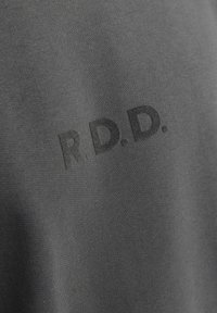 Šedá bavlněná tkanina s hladkou strukturou, na které jsou výrazně zobrazeny zvýšené tmavé nápisy "R.D.D.". Žádné další vzory nebo akcenty.