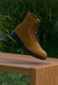 Brown suede ankle boot with round toe, lace-up design, and black rubber sole, set against a wooden surface with green grass background.