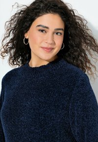 Navyblauw sweater van een gestructureerde, zachte stof. Heeft een ribbelkraag en lange mouwen, gedragen door een persoon met krullend haar.