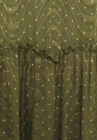 Olivově zelená tkanina s vlnitým vzorem a bílými puntíky, s volánkovými detaily v pase. Měkká textura s jemným leskem.